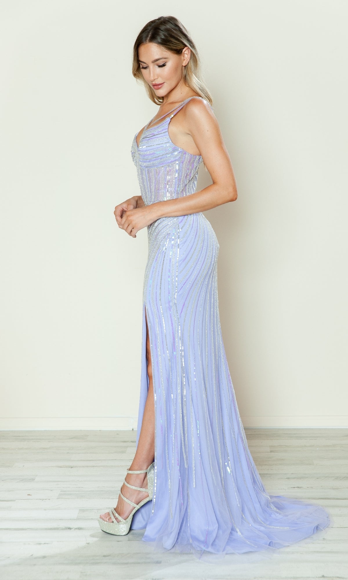 A woman stands in profile wearing the Sheer-Bodice Long Sequin Prom Dress (Poly USA 9664), a light purple, floor-length gown with sequins, side slit, and white high-heeled shoes against a plain light background.